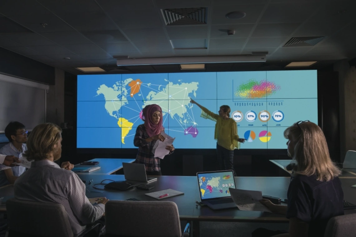People in a conference room listening to a presentation with geographical data projected on a screen.