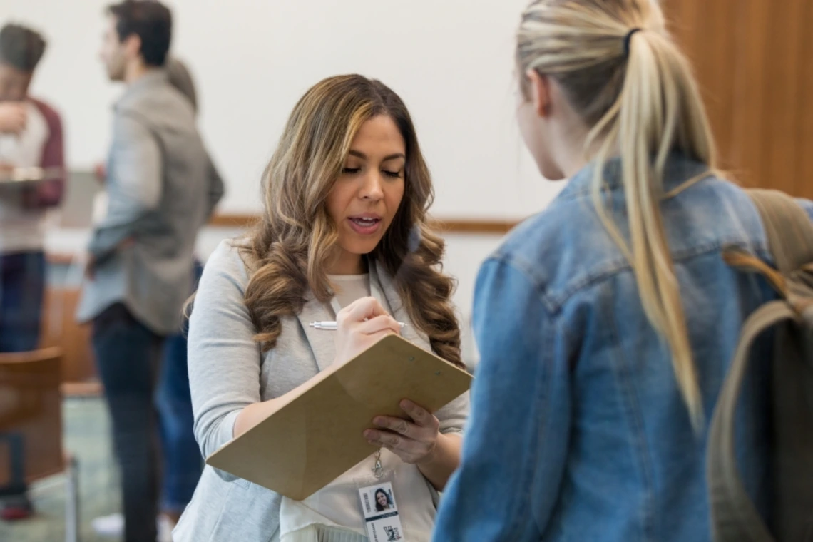 A healthcare worker writing notes on a clipboard while talking to another woman