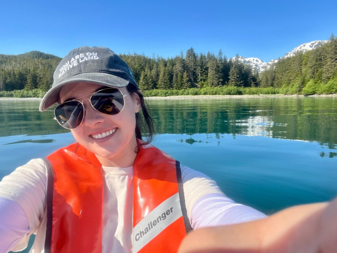 Halley taking a selfie while in the middle of the lake. They are wearing a life jacket and smiling. The lake is very clue and has pine trees surrounding its edges.