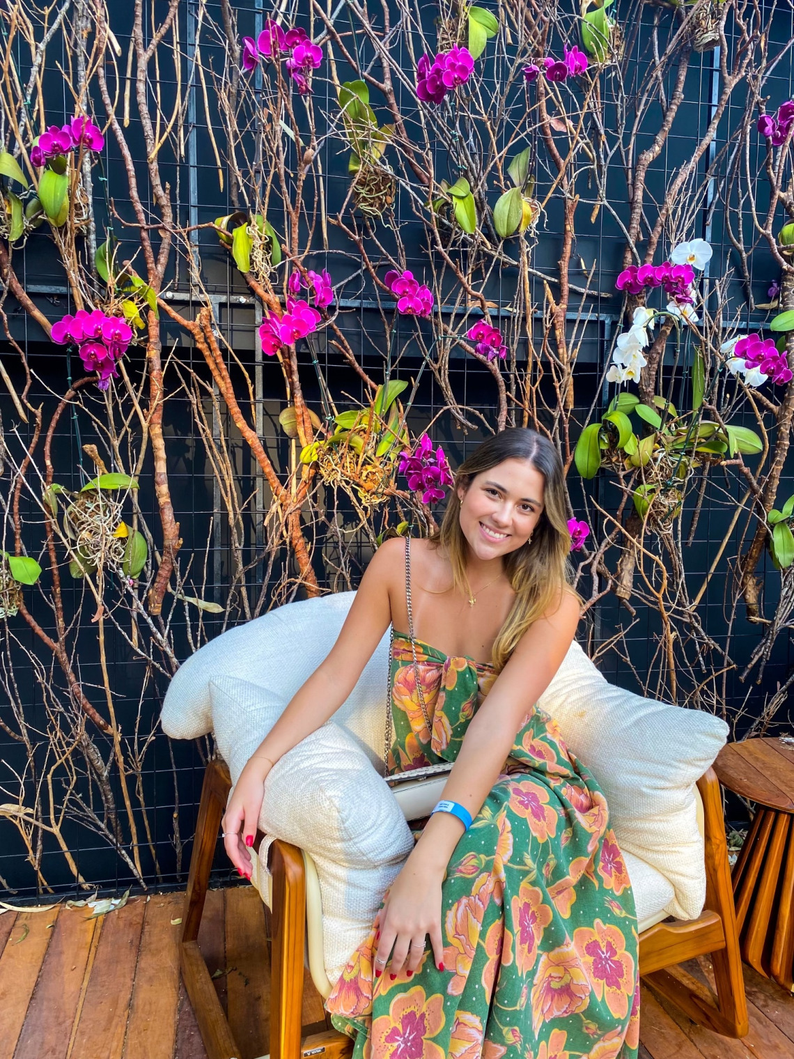 Maria smiling at the camera wearing a floral dress while sitting on a comfy chair. There are pretty vines with flowers in the background.